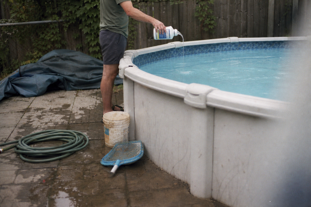 Person pours pool treatment from a bottle into an above-ground backyard swimming pool.